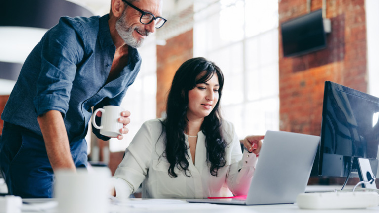 Two people working together. One in front of a laptop with colleague standing behind helping